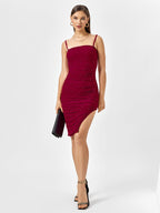 Woman wearing a red embellished ruched midi dress with spaghetti straps and an asymmetrical hem, holding a black clutch.