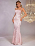 A woman poses in a light pink satin jacquard mermaid formal gown with a square neckline and adjustable tie straps.