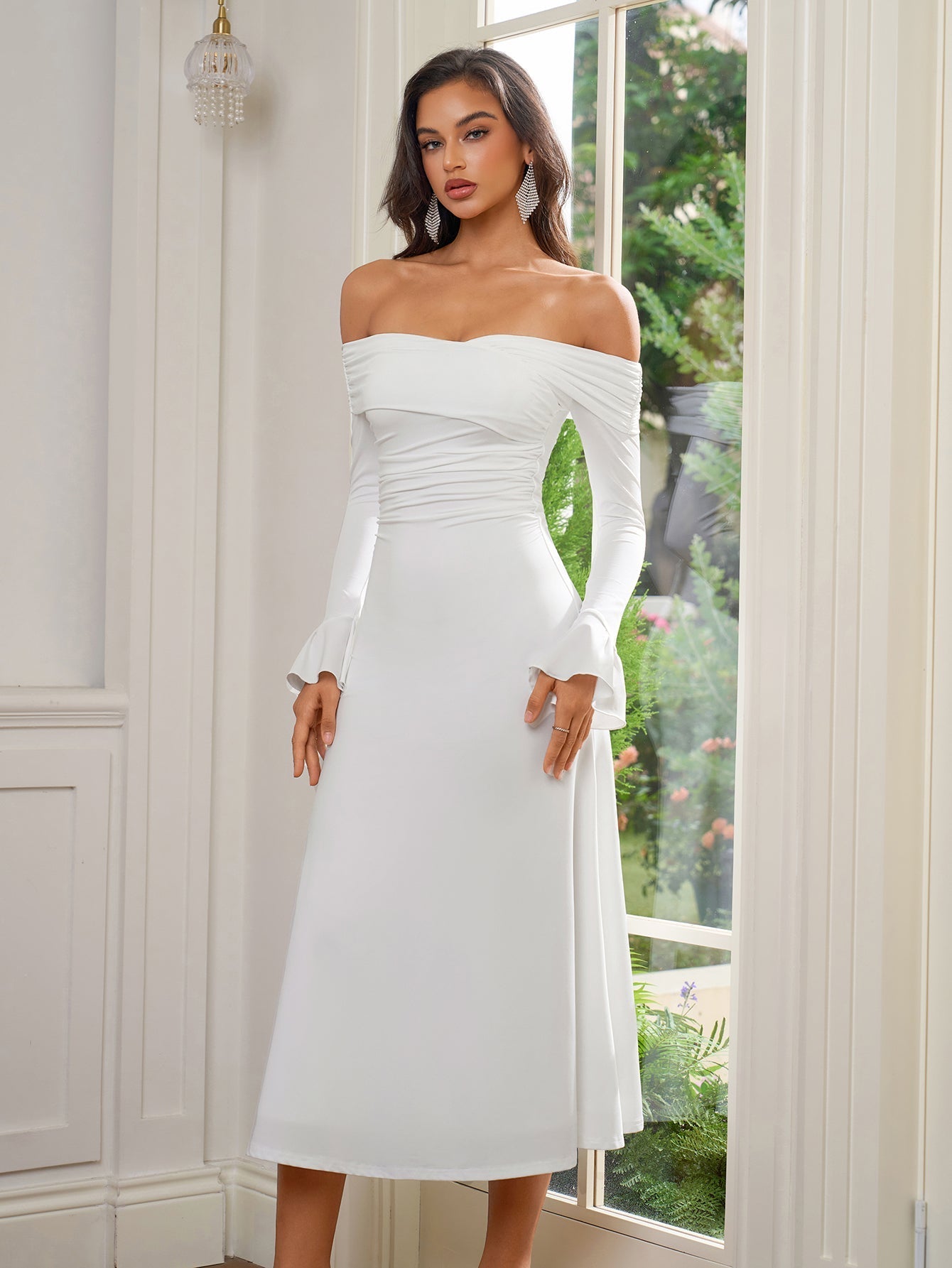 Full-length shot of a woman in a white off-the-shoulder midi dress, standing by a window.