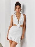 Woman leaning against a wall wearing a white sleeveless mini dress with a deep V-neck, ruffle trim, and cut-out details