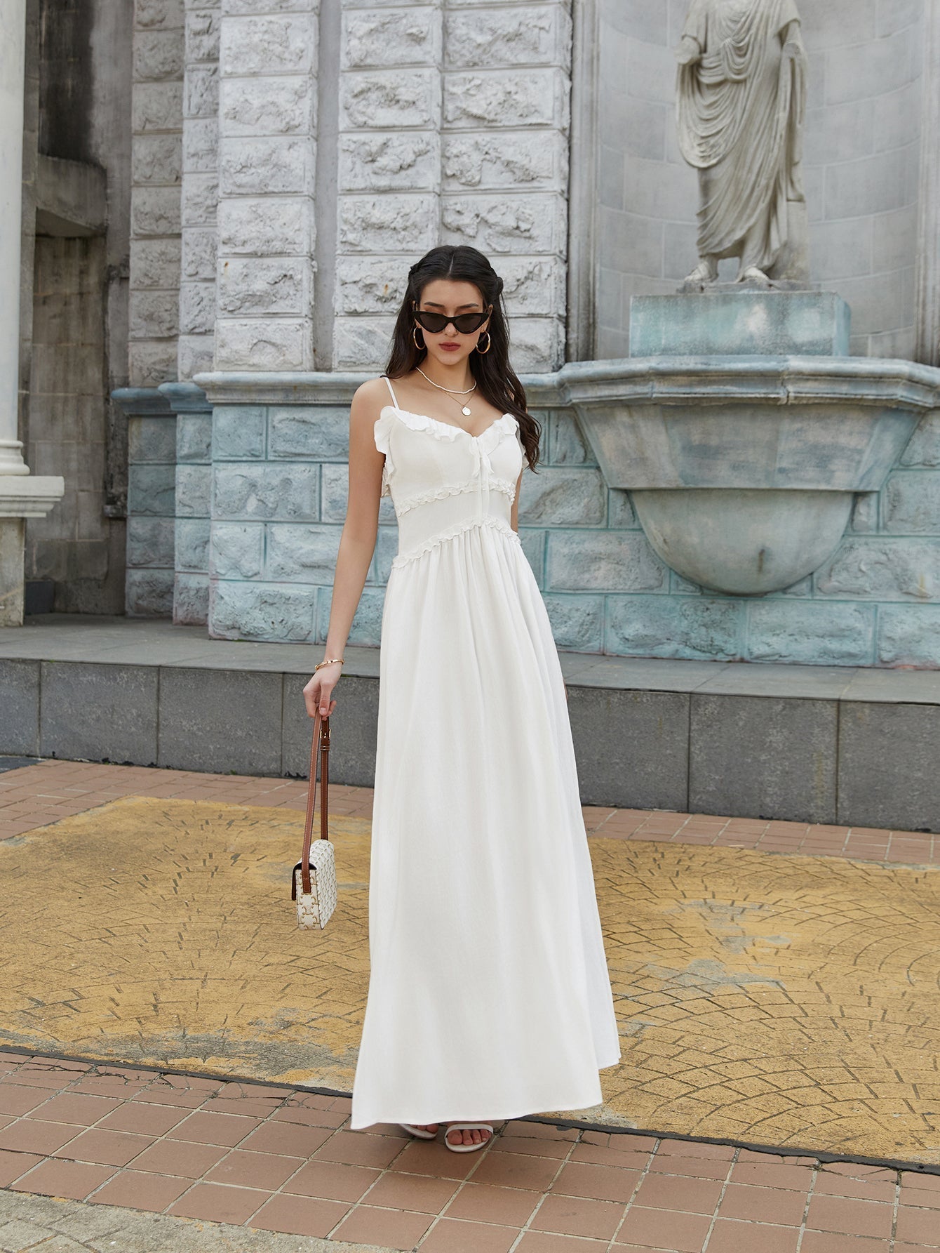 Woman in a white maxi dress with ruffled trim and spaghetti straps holding a small handbag.
