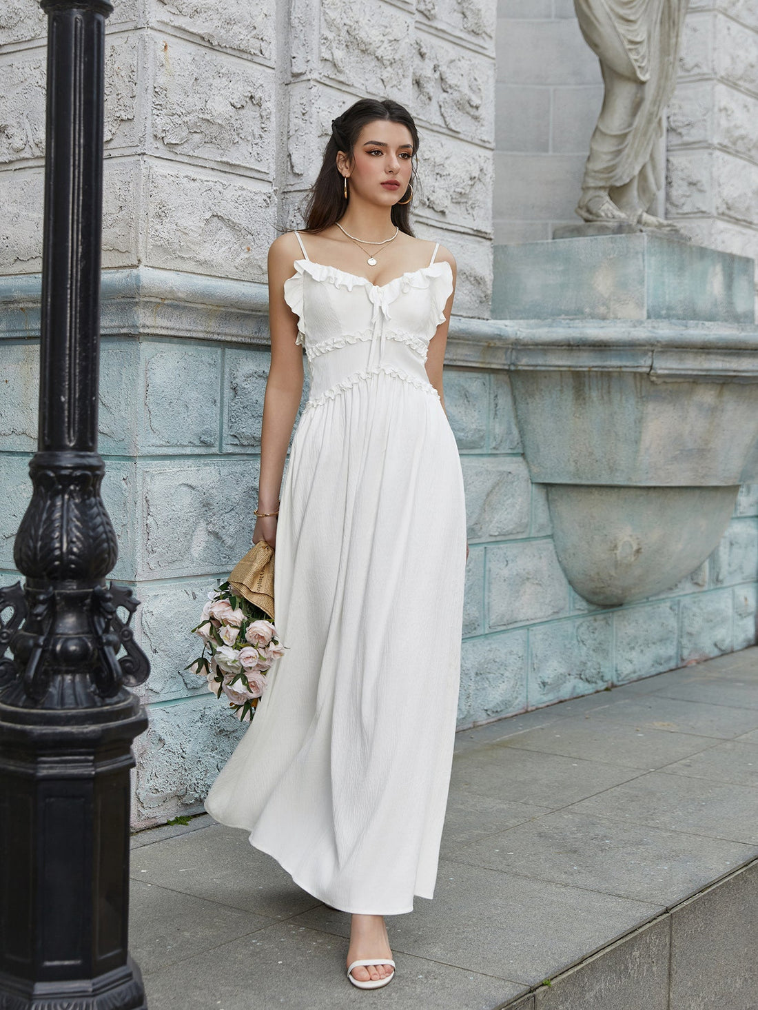 Woman in a white maxi dress with ruffled trim and spaghetti straps holding a bouquet of flowers.