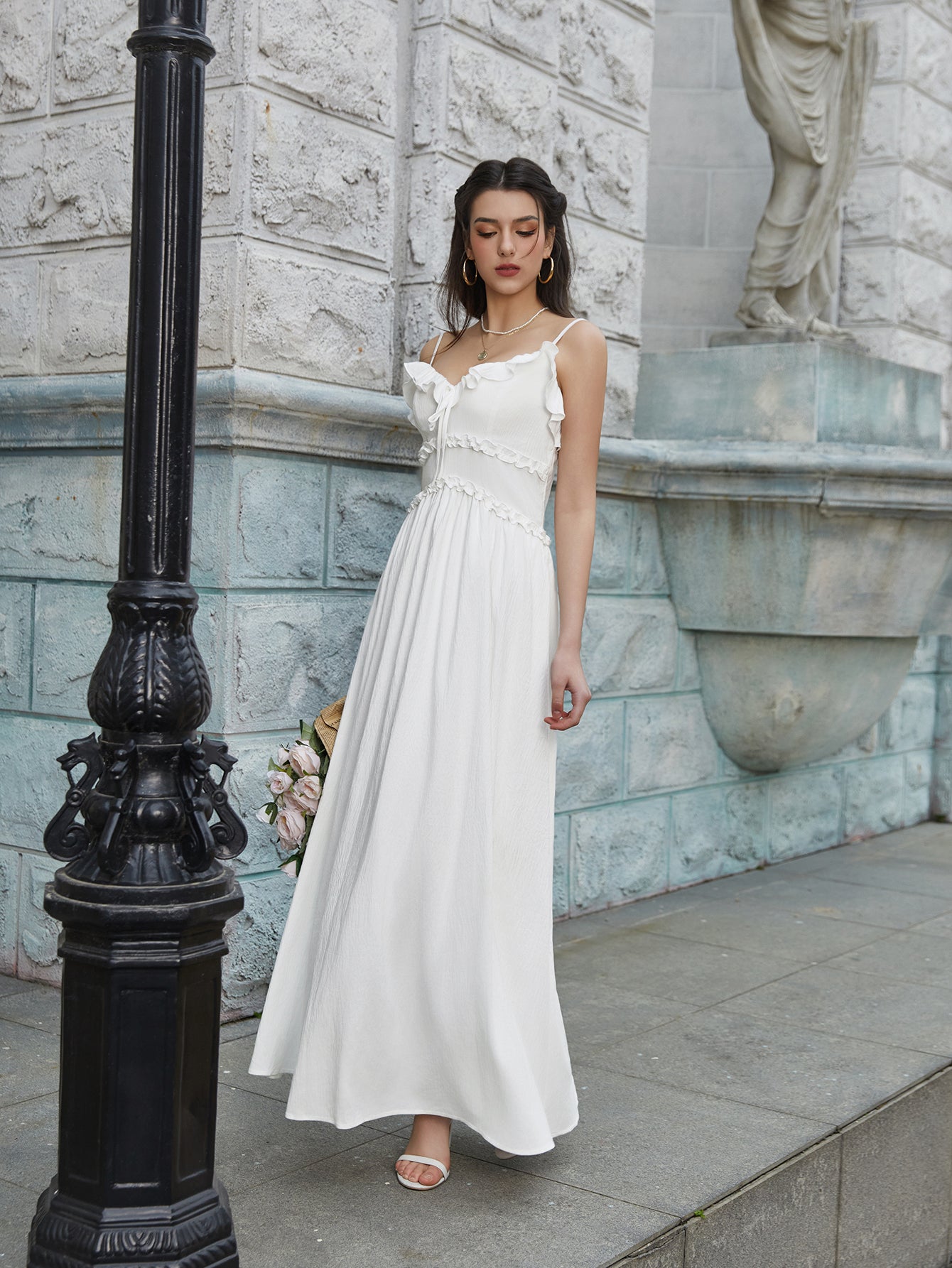Woman in a white maxi dress with ruffled trim and spaghetti straps standing by a lamppost.