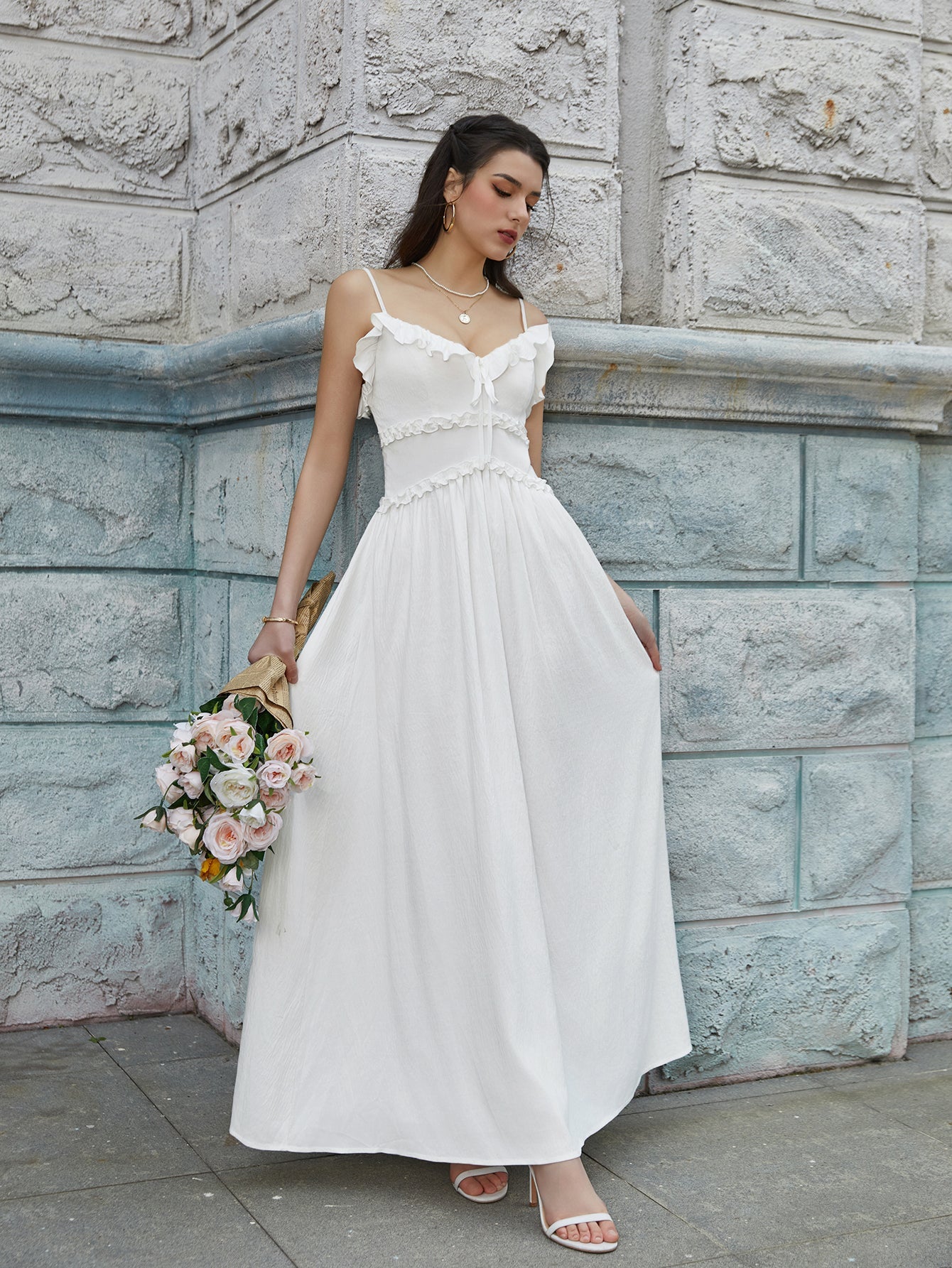 Woman in a white maxi dress with ruffled trim and spaghetti straps posing with flowers.