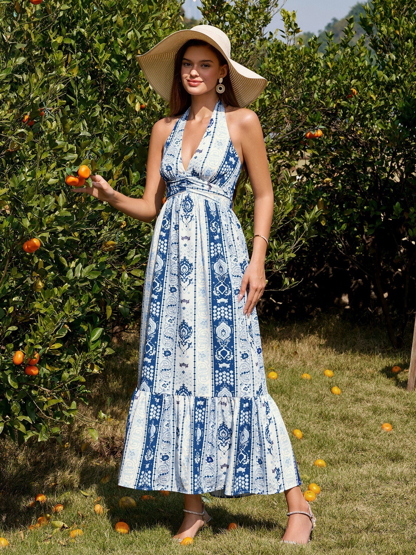 Woman wearing a blue and white patterned maxi dress, halter neckline, and a wide-brimmed straw hat, holding an orange