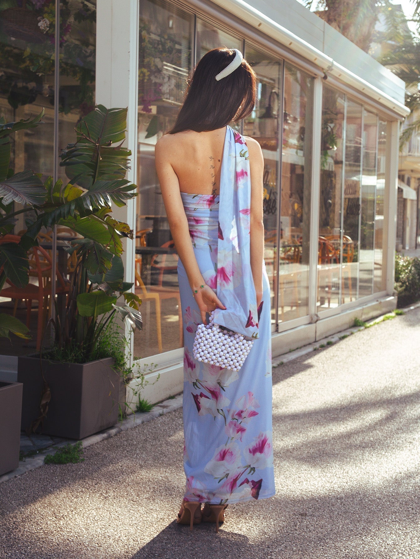 Back view of a woman in a light blue floral print maxi dress with a halter neckline and a draped scarf-like detail.