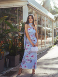 Full body shot of a woman in a light blue floral print maxi dress with a halter neckline and draped accents, standing outdoors.