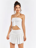 Front view of a woman in a white halter crop top with ruched detailing and an asymmetrical hem, and a white tiered mini skirt with a ruffled waistband.
