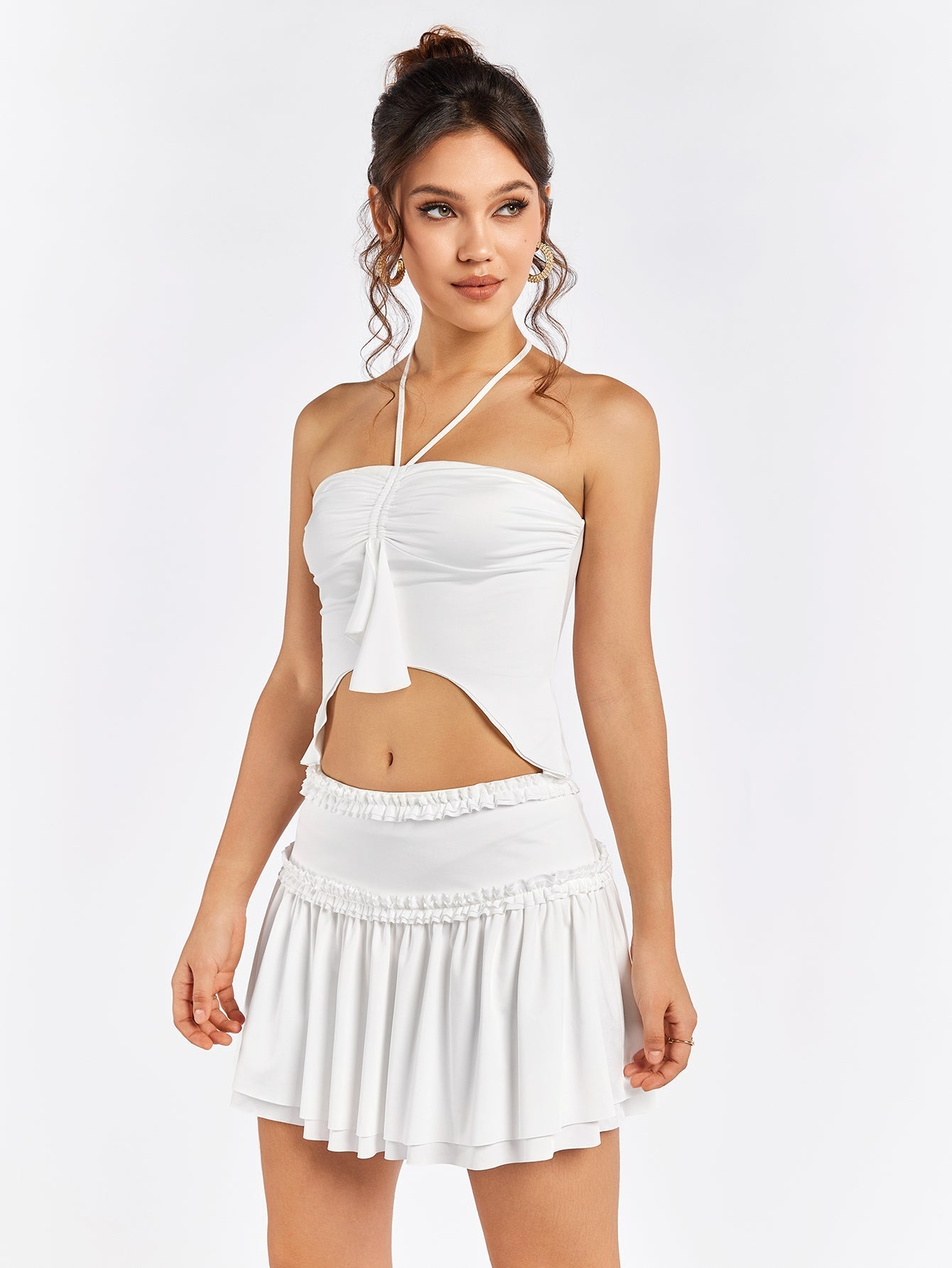 Front view of a woman in a white halter crop top with ruched detailing and an asymmetrical hem, and a white tiered mini skirt with a ruffled waistband.