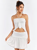 Woman posing in a white halter crop top with ruched detailing and an asymmetrical hem, and a white tiered mini skirt with a ruffled waistband.