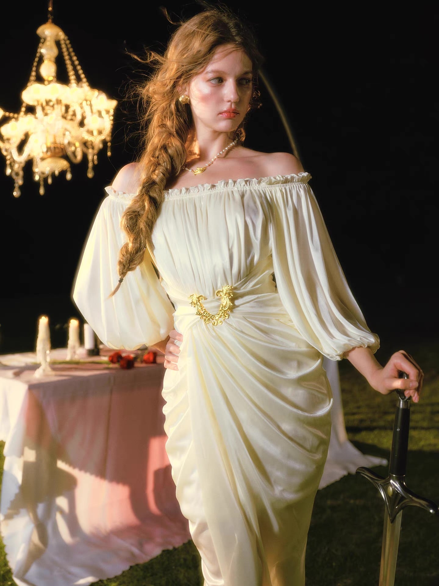 Full body shot of a woman in an elegant cream satin dress, holding a sword, with a chandelier and table setting in the background.