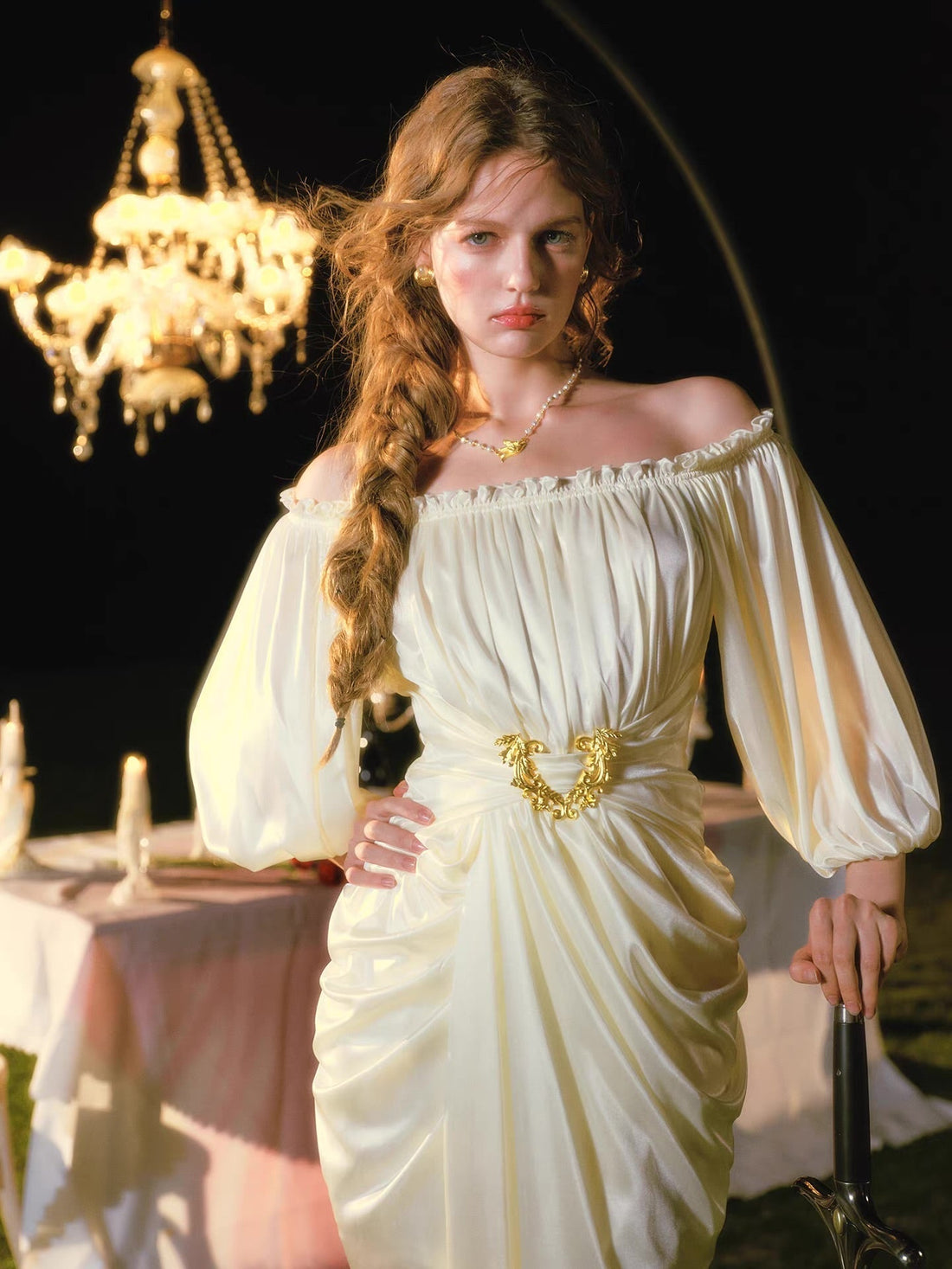 Woman in a cream off-the-shoulder satin dress with ruched detailing and a gold belt buckle, standing outdoors at night with a sword.
