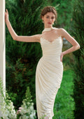Woman in a cream-colored silk satin mermaid gown with ruched detailing, leaning against a white pillar outdoors.