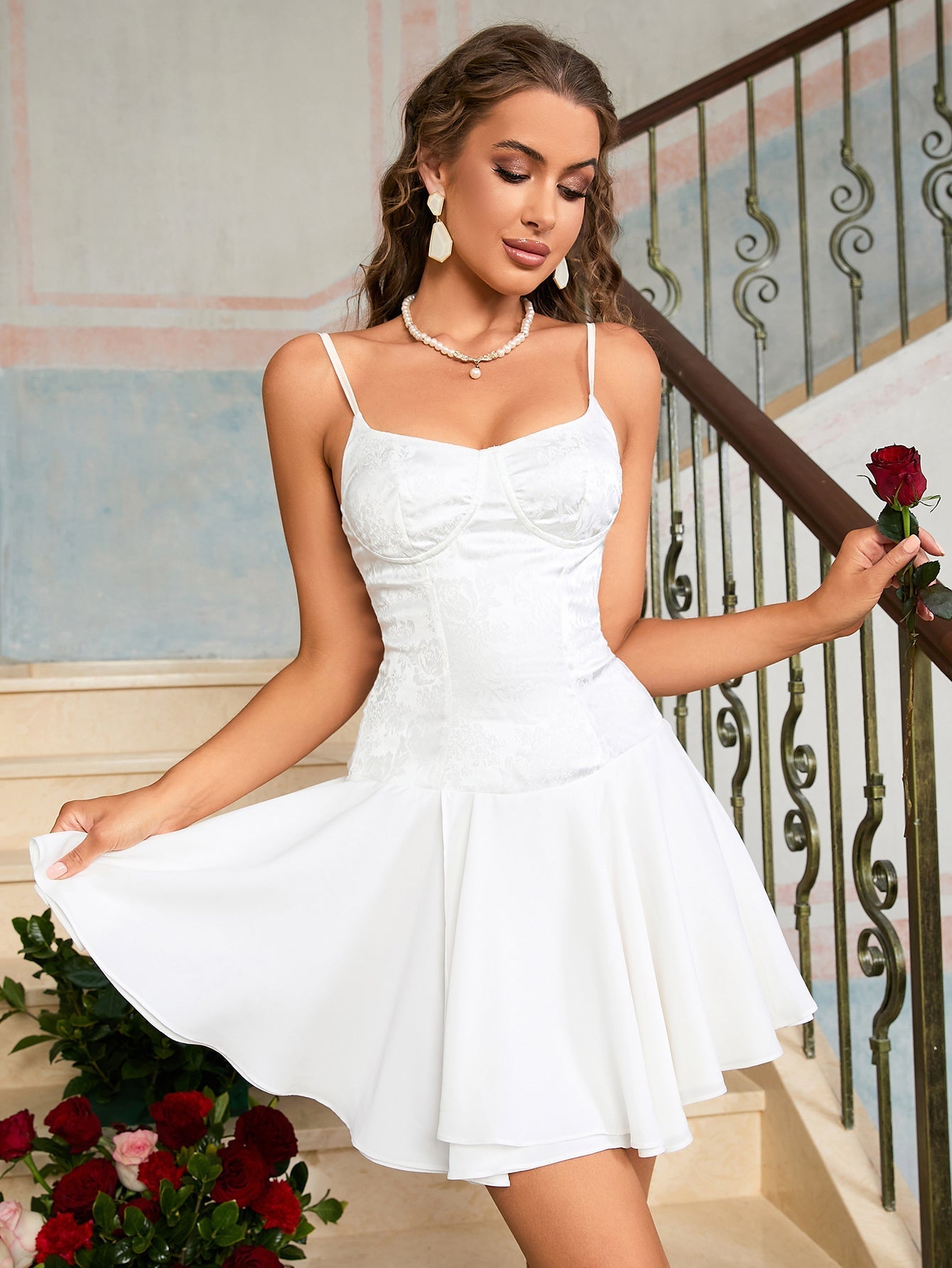 Woman holding a red rose, wearing a white satin dress with lace overlay and a pleated skirt.