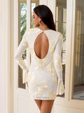 Back view of a woman in a white long-sleeve mini dress with a dramatic open back and floral detailing.