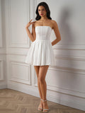 Full body shot of a woman in a white strapless embroidered dress with a pleated skirt, standing with her hand on her chest.