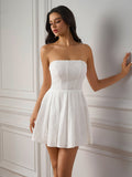 Woman in a white strapless embroidered dress with a pleated skirt, leaning against a wall.