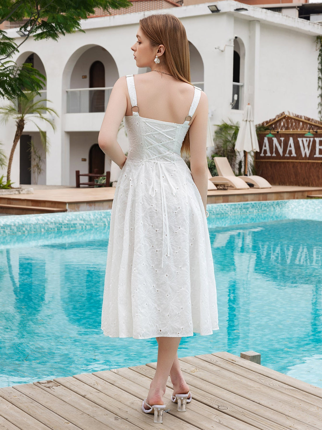 Woman wearing a white eyelet embroidered midi dress with V-neck and adjustable straps, standing on a wooden deck by a pool.