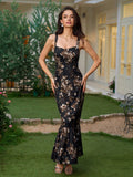 Full length view of a woman in a black floral print maxi dress with a square neckline and ruffled fishtail hem, hands on hips, standing in a garden.