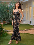 Woman in a black floral print maxi dress with a square neckline and ruffled fishtail hem, standing with one leg forward in a garden.