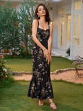 Woman in a black floral print maxi dress with a square neckline and ruffled fishtail hem, posing in a garden.