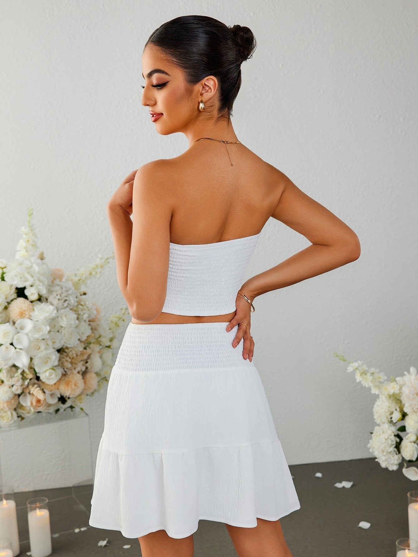 Rear view of a woman in a white strapless cropped top and a white tiered mini skirt with a smocked waistband.