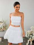 Woman wearing a white strapless cropped top with a tie front and eyelet detail, paired with a white tiered mini skirt with a smocked waistband.