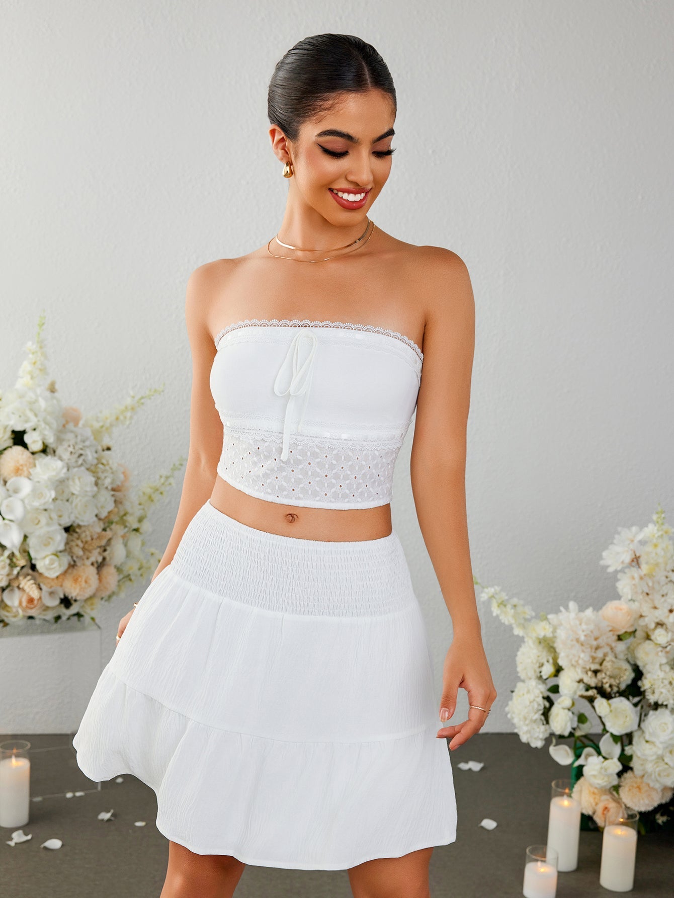 Woman wearing a white strapless cropped top with a tie front and eyelet detail, paired with a white tiered mini skirt with a smocked waistband.