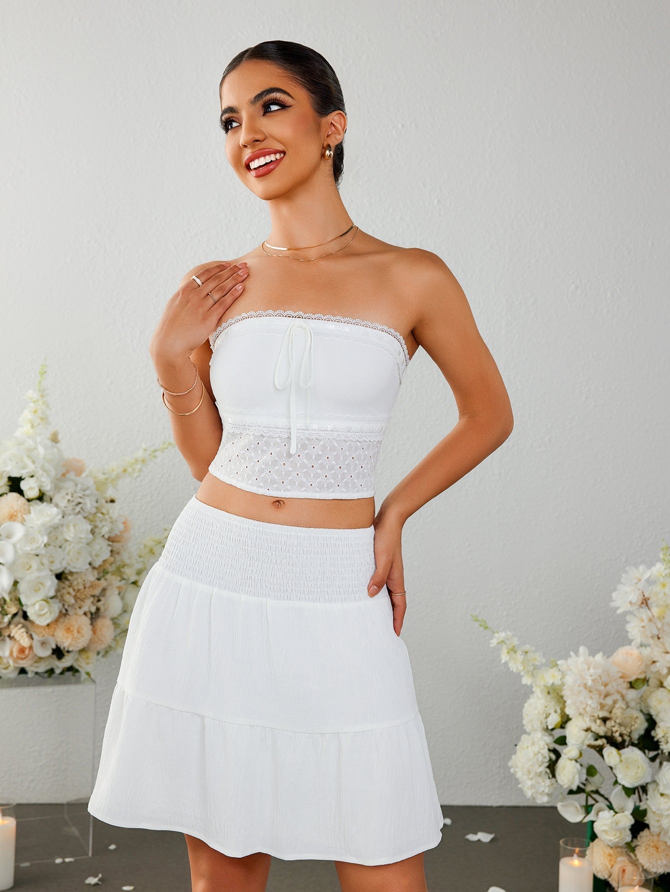 Woman posing in a white strapless cropped top with a tie front and eyelet detail, and a white tiered mini skirt with a smocked waistband.