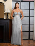 Woman in a silver metallic lurex A-line evening gown with a sweetheart neckline, cut-out waist, and high slit, with hands behind her back, posing by a fireplace.