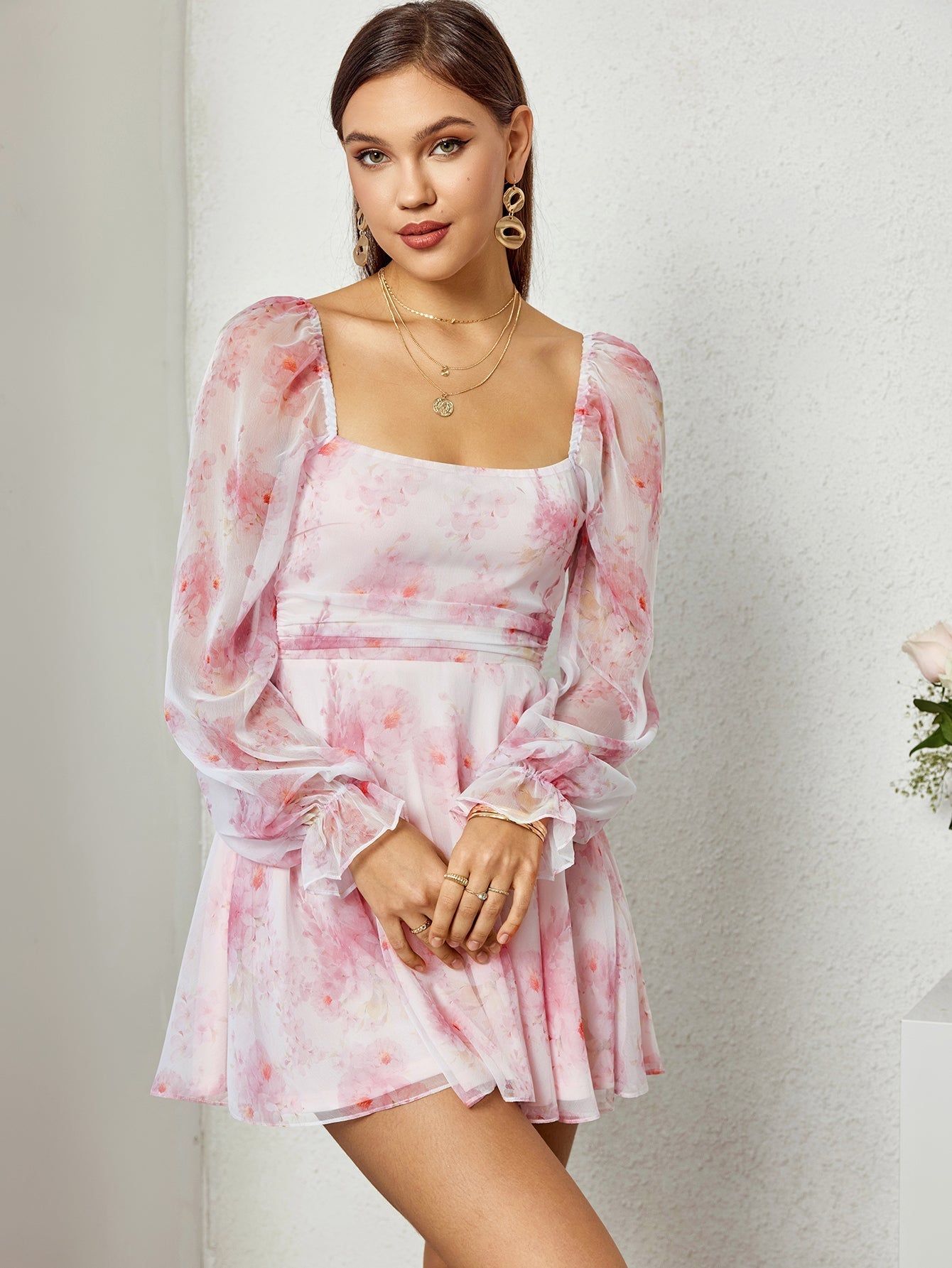 Woman in a pink floral print dress with a square neckline and long sleeves, standing against a textured wall.