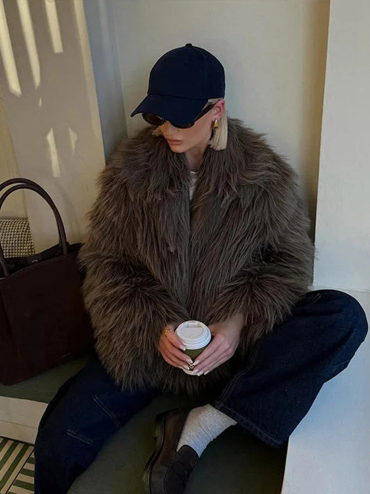Woman wearing a brown shaggy faux fur jacket, dark wash wide-leg jeans, and a navy baseball cap, sitting down and holding a coffee cup.