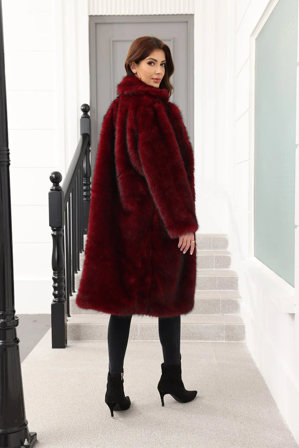 Rear view of a woman wearing a long, dark red faux fur coat with a relaxed fit, standing on stairs.