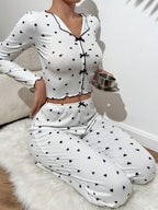 Woman wearing a white long-sleeved pajama top and pants set with a black heart print, holding a small metal cup and saucer, seated on a white fluffy rug.