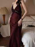 Woman in a burgundy satin slip dress with lace detailing, standing next to a bed.