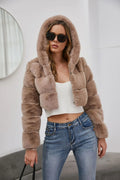 Woman wearing a cropped taupe faux fur hooded jacket with a white crop top and blue jeans.