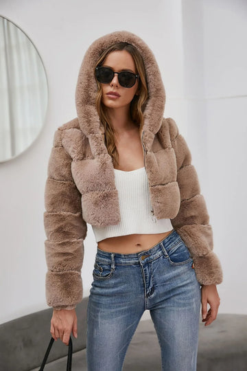 Woman wearing a cropped taupe faux fur hooded jacket with a white crop top and blue jeans.