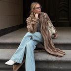 Woman wearing a long brown faux fur coat, white t-shirt, and light blue wide-leg jeans, sitting on steps and resting her chin on her hand.