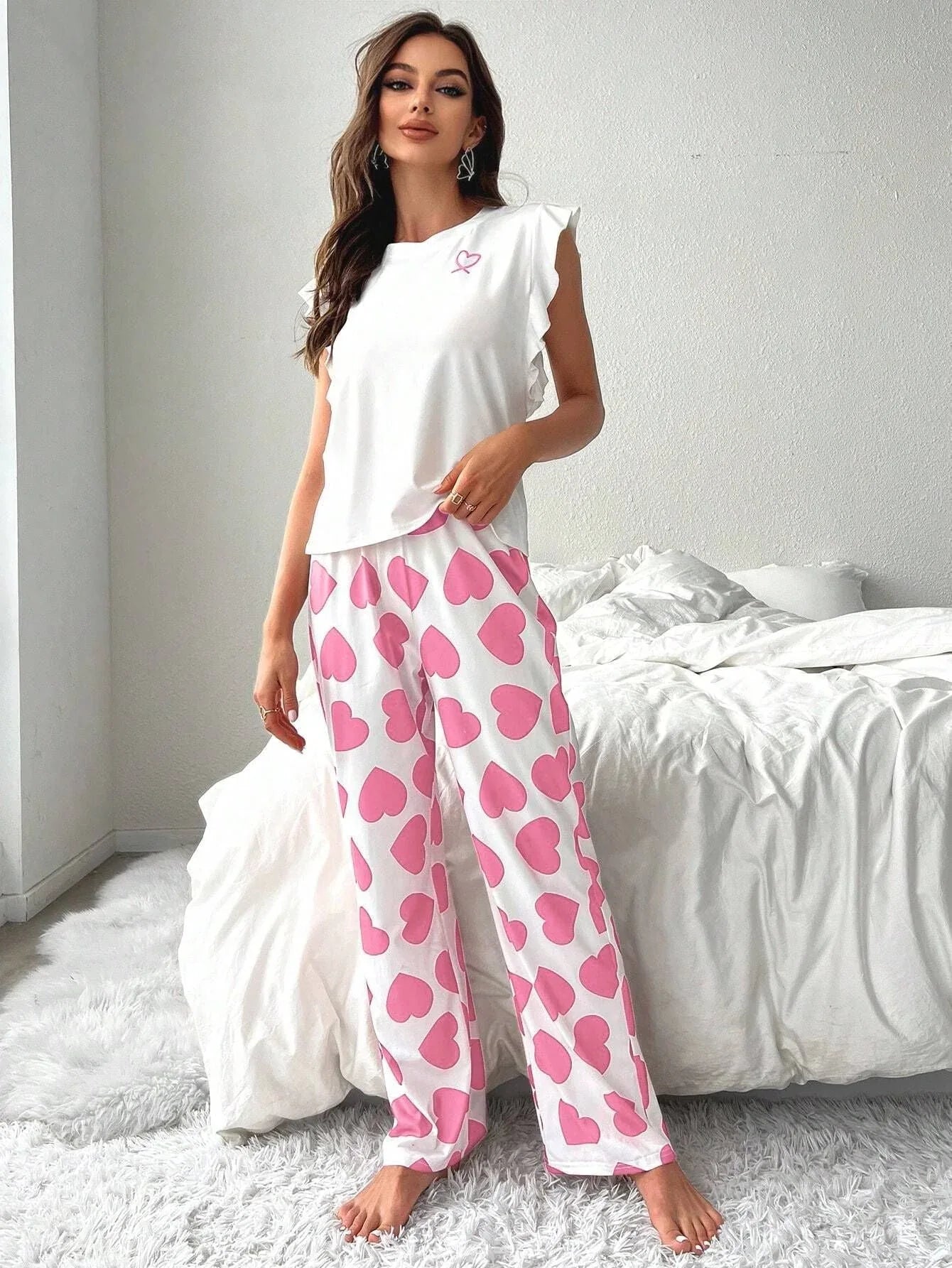 Woman wearing white top with ruffled sleeves and pink heart print pajamas standing in a bedroom Main image