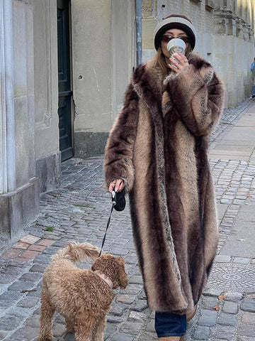 Woman wearing a long, striped faux fur coat and a knit hat, walking a dog.