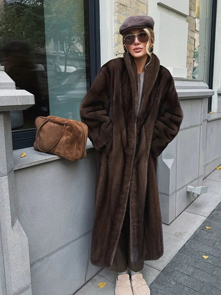 Full body shot of a woman in a long brown faux fur coat, taupe knit sweater, taupe trousers, taupe beret, sunglasses, and tan slip-on shoes, with a brown suede handbag resting on a ledge.