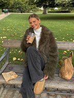 Woman wearing a green faux fur jacket, white turtleneck, and dark jeans sitting on a park bench with a coffee.