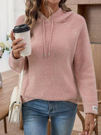 Woman holding a coffee cup and a handbag, wearing a pink hooded knit sweater and blue denim jeans