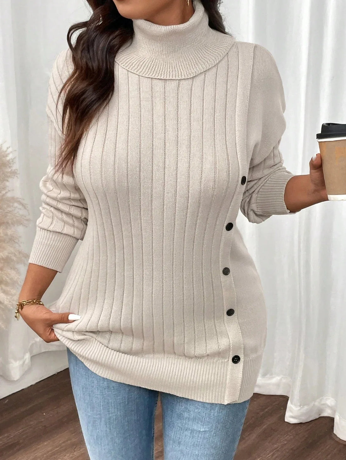 Woman wearing a cream ribbed knit turtleneck tunic sweater with button detail on the side, paired with blue jeans.
