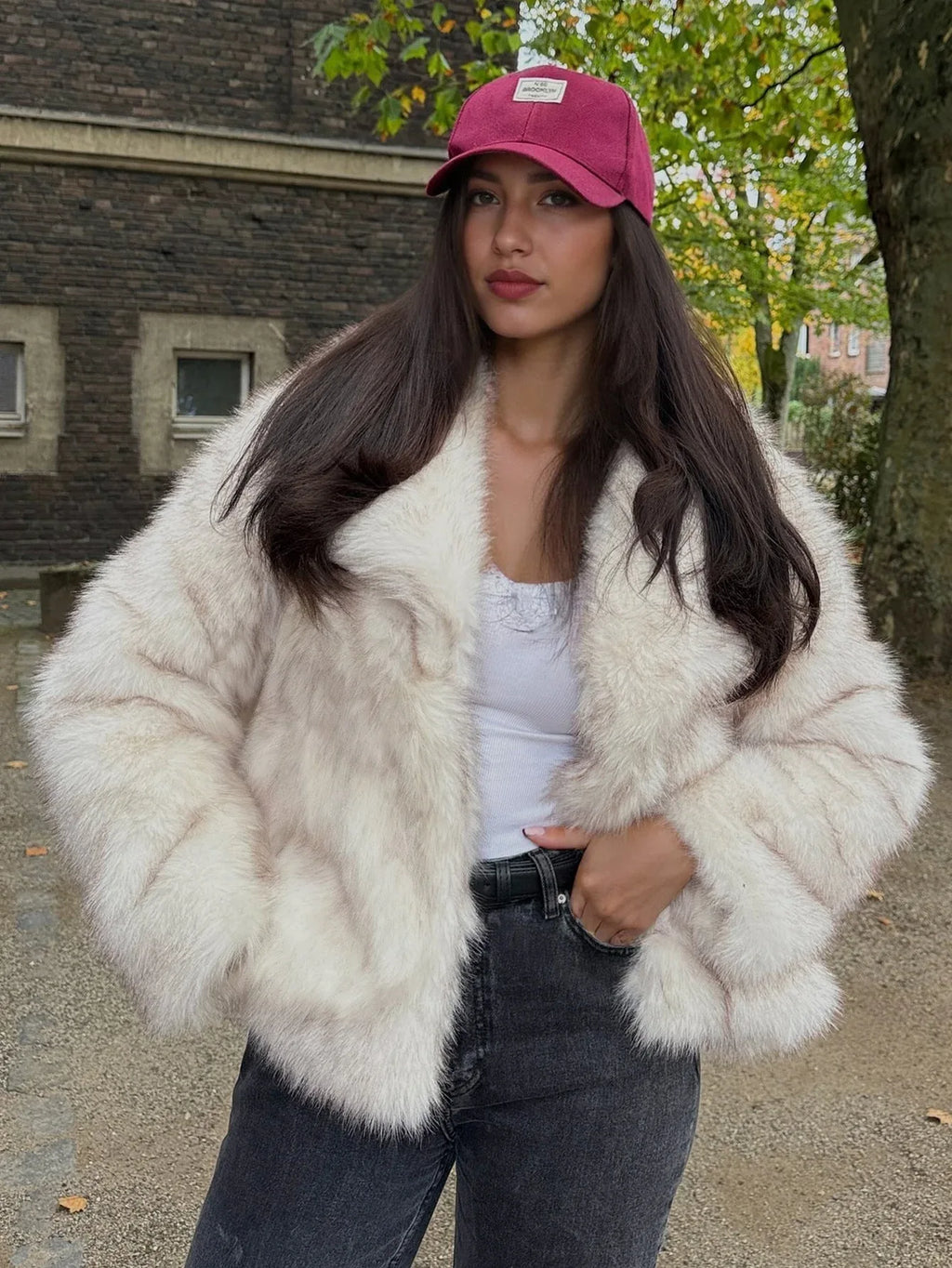 Fashionable woman wearing a cream faux fur jacket, dark jeans, and a pink baseball cap, looking at the camera.
