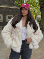 Fashionable woman wearing a cream faux fur jacket, dark jeans, and a pink baseball cap, looking at the camera.
