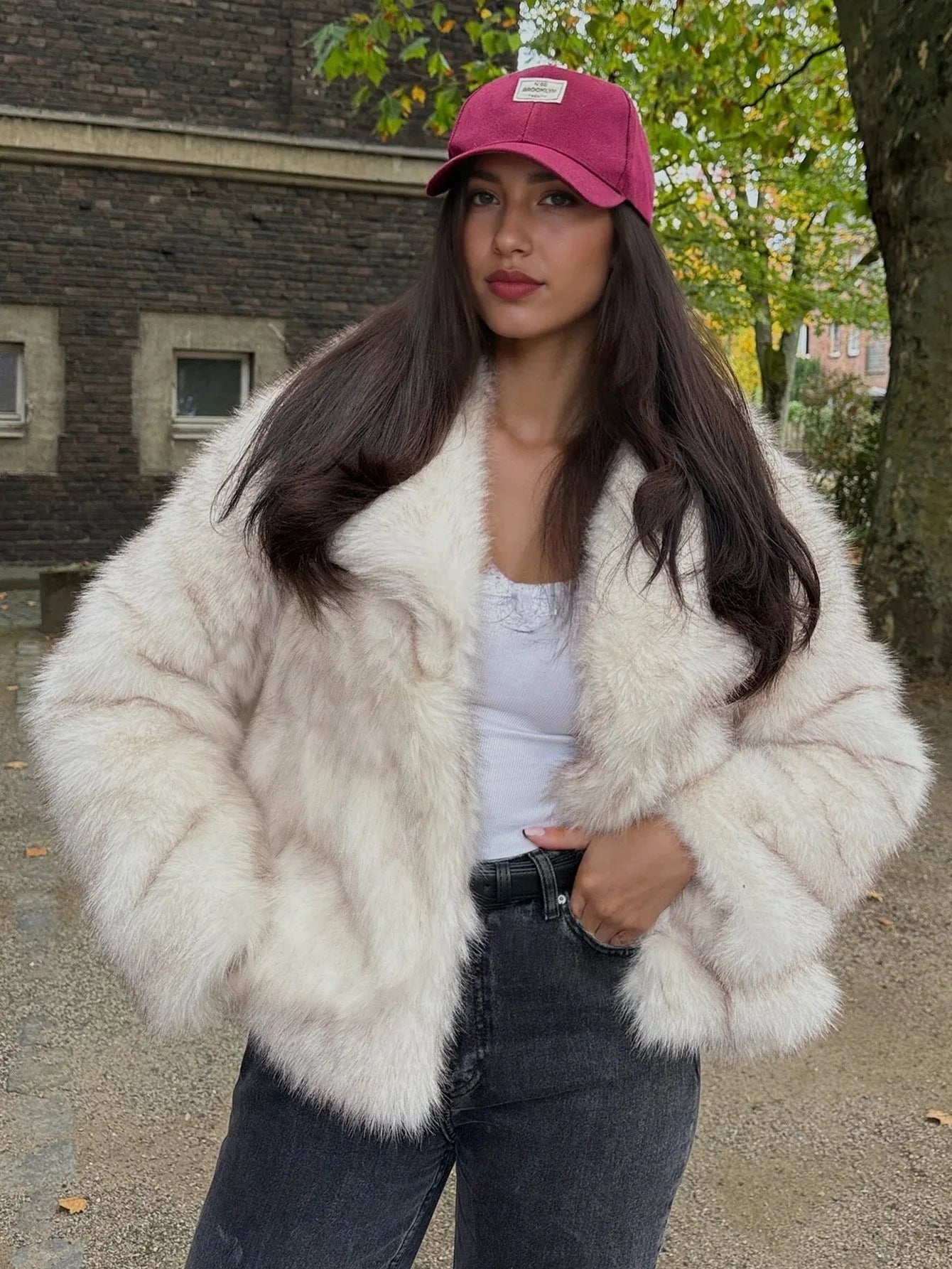 Fashionable woman wearing a cream faux fur jacket, dark jeans, and a pink baseball cap, looking at the camera.