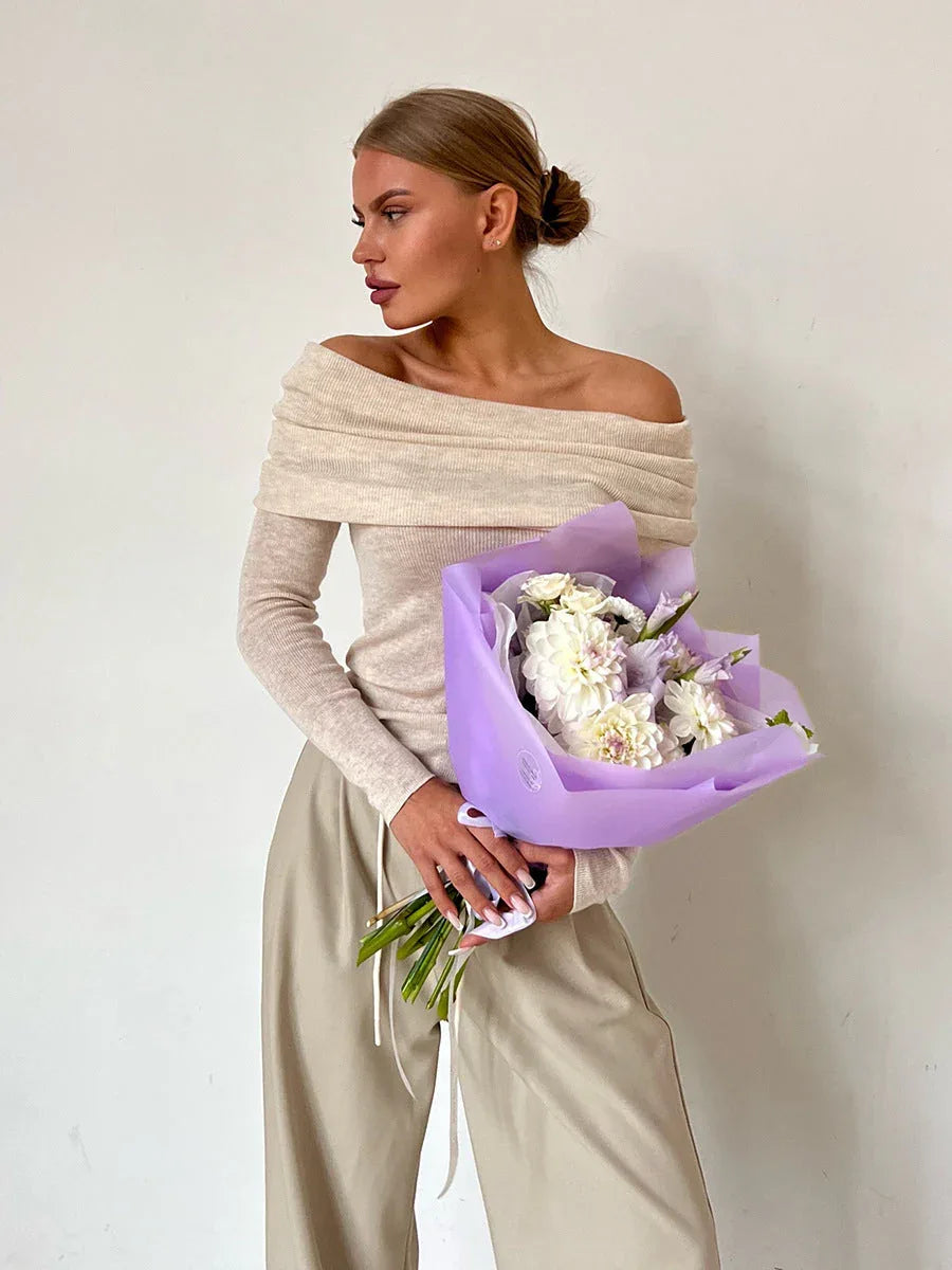 Woman holding a bouquet of white and purple flowers while wearing a beige off-the-shoulder sweater.