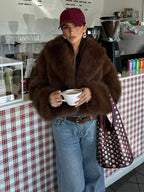 Woman wearing a brown faux fur jacket, blue jeans, and a red baseball cap holding a coffee cup