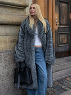 Woman standing outdoors wearing a long grey faux fur coat with a lapel collar and full length sleeves, styled with blue jeans.